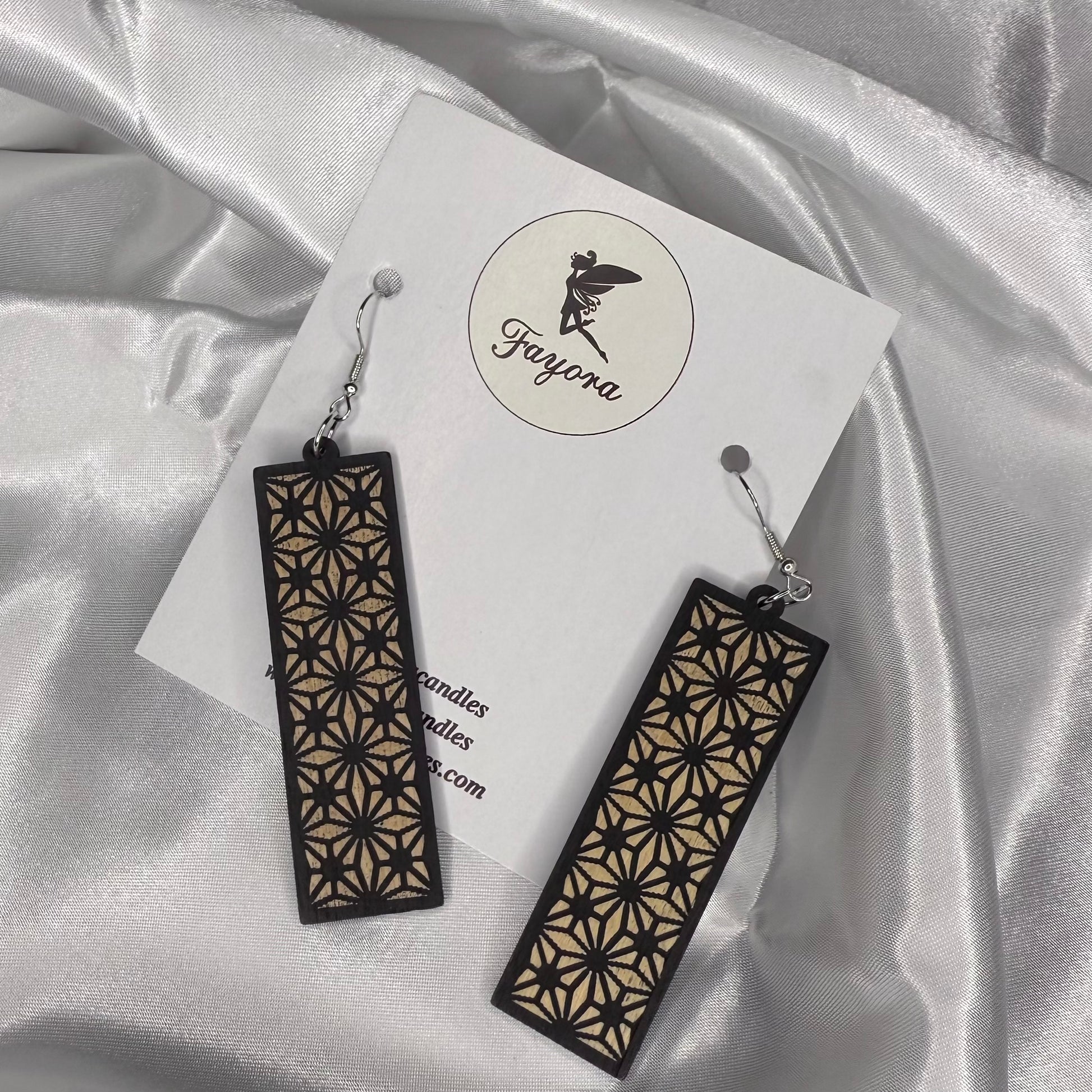 Pair of black and gold geometric earrings on a white card with 'Fayora' branding, against a satin fabric background.